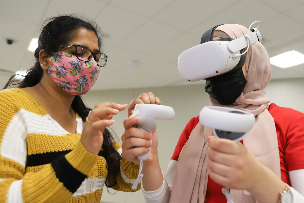 Woman on the left instructing woman on the right how to use VR headset and joysticks
